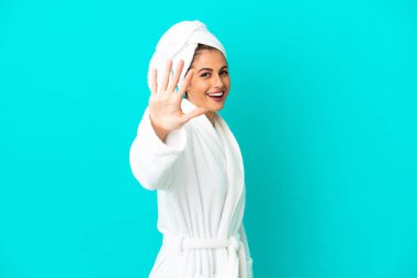 İzole edilmiş mavi arka planda bornozlu genç bir kadın parmaklarıyla beşi sayıyor.