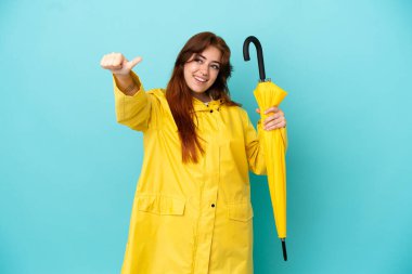 Mavi arka planda şemsiyesi olan kızıl saçlı kadın baş parmak hareketi yapıyor.