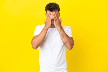 Beyaz tenli yakışıklı adam sarı arka planda izole edilmiş gözlerini elleriyle kapatıyor.