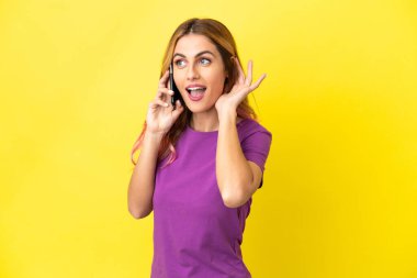 İzole edilmiş sarı arka planda cep telefonu kullanan genç bir kadın kulağına elini sürerek bir şeyler dinliyor.