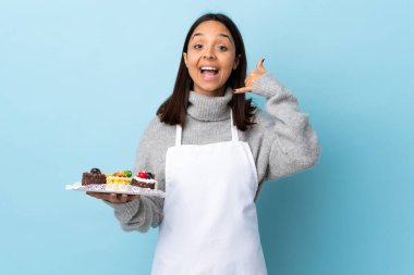 Pasta şefi, izole edilmiş mavi arka planda büyük bir pasta tutuyor ve telefon jesti yapıyor. Bana geri dönüş işareti ver..