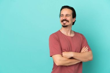 Mavi arka planda izole edilmiş, kolları bağlı ve mutlu genç bir beyaz adam.