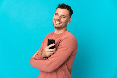 Mavi arka planda izole edilmiş genç beyaz bir adam cep telefonu tutuyor ve kollarını kavuşturuyor.