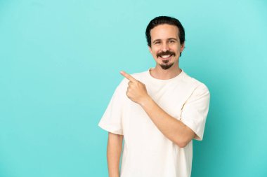 Mavi arka planda izole edilmiş genç beyaz adam bir ürünü sunmak için kenarı işaret ediyor.
