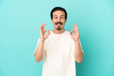 Mavi arka planda izole edilmiş beyaz bir adam iki eliyle tamam işareti gösteriyor.