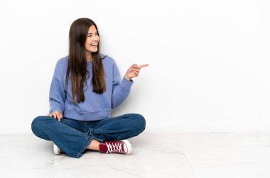 Genç bir kadın yerde oturmuş, parmağını kenarı işaret ediyor ve bir ürün sunuyor.