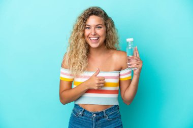 Mavi arka planda izole edilmiş su şişesi olan genç sarışın bir kadın baş parmak işareti yapıyor.