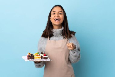 Pasta şefi elinde büyük bir pasta tutuyor mavi arka planda şaşırmış ve ön tarafı işaret ediyor..