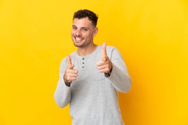 Sarı arka planda izole edilmiş beyaz, yakışıklı genç bir adam ön tarafı işaret ediyor ve gülümsüyor.