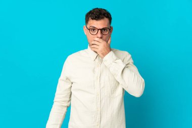 Beyaz tenli yakışıklı bir adam mavi arka planda izole edilmiş. Ağzını eliyle kapatıyor.