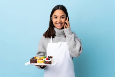 Pasta şefi, mavi arka planda büyük bir pasta tutuyor ve cep telefonuyla sohbet ediyor..