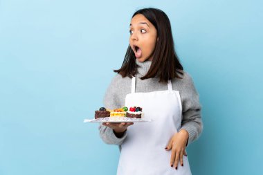 Pasta şefi mavi arka planda büyük bir pasta tutuyor. Yan tarafa bakarken sürpriz jest yapıyor..