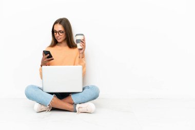 Dizüstü bilgisayarı olan genç bir kadın elinde kahve ve cep telefonu ile yerde oturuyor.