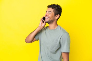 Genç ve yakışıklı Brezilyalı bir adam sarı arka planda izole edilmiş cep telefonuyla sohbet ediyor.