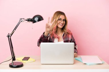 Pembe arka planda dizüstü bilgisayarı olan genç bir bayan öğrenci mutlu ve parmaklarıyla üçe kadar sayıyor.