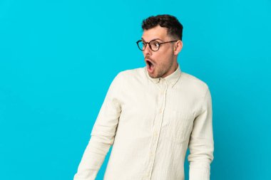 Genç, beyaz, yakışıklı bir adam mavi arka planda izole edilmiş. Yan tarafa bakarken sürpriz jest yapıyor.