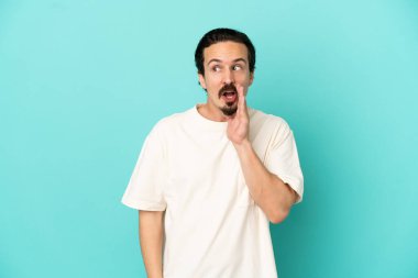 Mavi arka planda izole edilmiş genç beyaz adam yan tarafa bakarken sürpriz bir hareketle bir şeyler fısıldıyor.