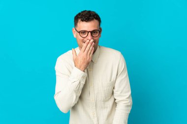 Genç, beyaz, yakışıklı bir adam mavi arka planda izole edilmiş mutlu ve gülümseyen bir ağızla elini kapatıyor.