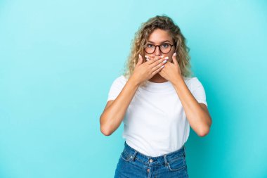Kıvırcık saçlı kız, mavi arka planda izole edilmiş, ağzı elleriyle kapatılmış.