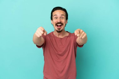 Mavi arka planda izole edilmiş genç beyaz adam şaşırmış ve ön tarafı işaret ediyor.