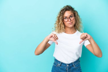 Mavi arka planda izole edilmiş kıvırcık saçlı kız baş parmağını iki eliyle aşağı gösteriyor.