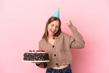 Elinde doğum günü pastası tutan Litvanyalı genç bir kadın pembe arka planda zaferi kutluyor.