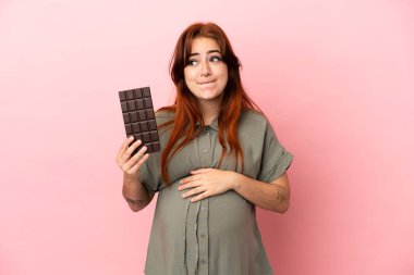 Genç kızıl saçlı beyaz kadın pembe arka planda izole edilmiş hamile ve elinde aç çikolatayla.