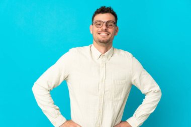 Beyaz tenli yakışıklı adam mavi arka planda izole edilmiş, kolları kalçasında ve gülümsüyor.