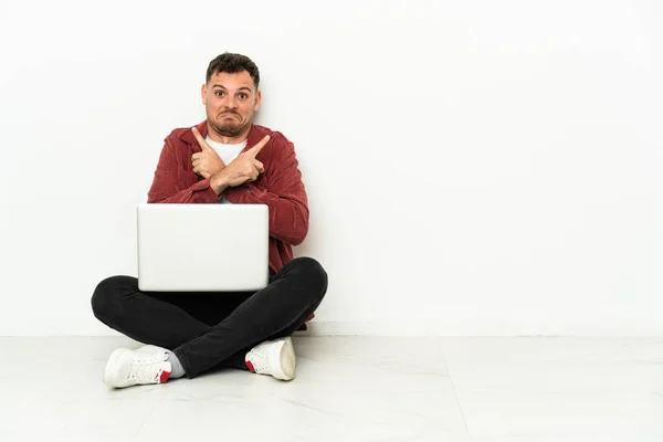 年轻英俊的高加索男子坐在地板上 膝上型电脑指向侧边 令人怀疑 — 图库照片