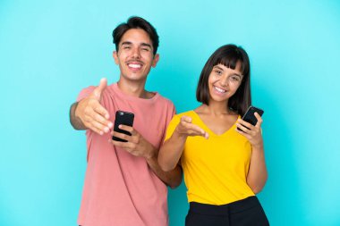 Mavi arka planda cep telefonu tutan genç melez çift iyi bir anlaşma için el sıkışıyor.
