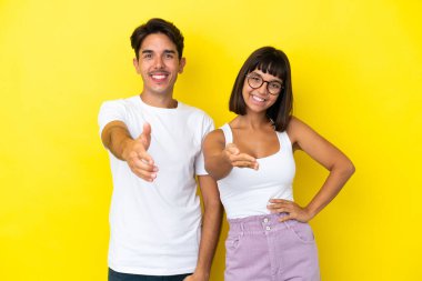 Genç melez çift sarı arka planda izole edilmiş iyi bir anlaşma için el sıkışıyorlar.