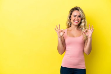 Sarı arka planda izole edilmiş genç Brezilyalı kadın iki eliyle tamam işareti gösteriyor.