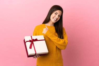 İzole edilmiş pembe arka planda elinde bir hediye tutan Brezilyalı genç kız baş parmak işareti yapıyor.