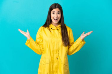 Yağmura dayanıklı ceket giyen Brezilyalı genç bir kız izole edilmiş mavi bir arka plan ve şok olmuş yüz ifadesi.