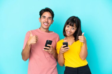 Mavi arka planda cep telefonu tutan melez bir çift iki eliyle baş parmak işareti yapıyor ve gülümsüyor.