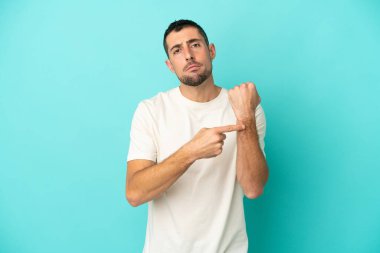 Mavi arka planda izole edilmiş yakışıklı beyaz adam geç kalma jesti yapıyor.