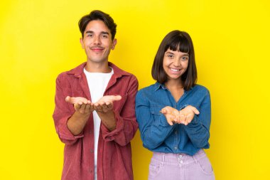 Genç melez çift sarı arka planda izole edilmiş. Avuçlarında bir ilan vermek için hayali bir alan tutuyorlar.