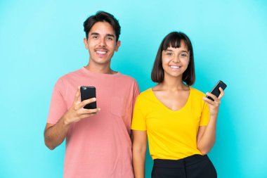 Mavi arka planda cep telefonu tutan genç melez çift kollarını kalçalarına dayayıp gülümsüyor.