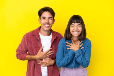 Sarı arka planda izole edilmiş genç melez çift ellerini göğsüne koyup gülümsüyor.