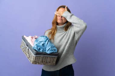 Orta yaşlı bir kadın, mor arka planda, elleri ve gözleri kaplayan bir çamaşır sepetini tutuyor. Bir şey görmek istemiyorum.