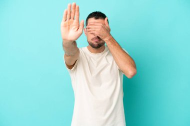 Mavi arka planda izole edilmiş yakışıklı beyaz adam jesti durdurup yüzünü kapatıyor.