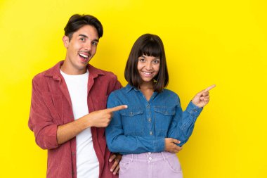 Sarı arka planda izole edilmiş genç ve melez bir çift parmak göstererek bir ürün sunuyor.