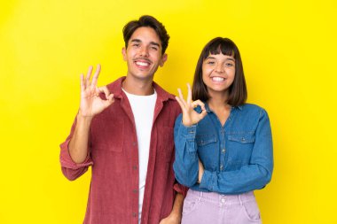 Sarı arka planda izole edilmiş genç melez çift parmaklarıyla tamam işareti gösteriyor.