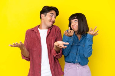 Genç melez çift sarı arka planda izole edilmiş. Omuzlarını kaldırırken önemsiz bir jest yapıyorlar.