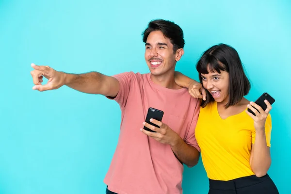 年轻的混血种族夫妇手持蓝色背景的手机 指向侧面展示产品 — 图库照片