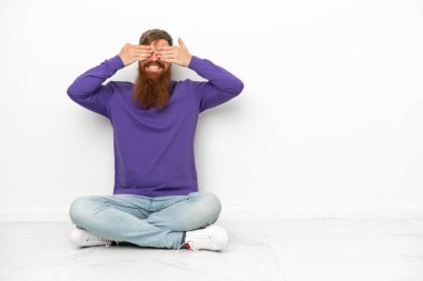 Beyaz arka planda izole bir şekilde yerde oturan Kafkasyalı genç adam elleriyle gözlerini kapatıyor.