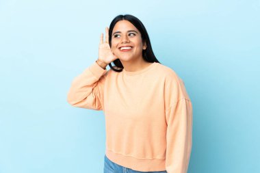 Mavi arka planda izole edilmiş genç Latin bir kadın. Kulağına elini sürerek bir şeyler dinliyor.