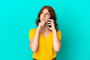 Cep telefonu kullanan genç kızılımsı kadın mavi arka planda izole edilmiş ve yukarı bakıyor.