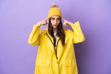 Mor arka planda izole edilmiş, yağmura dayanıklı bir ceket giyen beyaz bir kadın. Şüpheleri ve düşünceleri var.