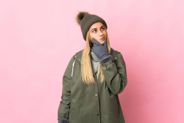 Pembe arka planda izole edilmiş, kış şapkalı genç bir kadın yukarı bakarken şüphe duyuyor.